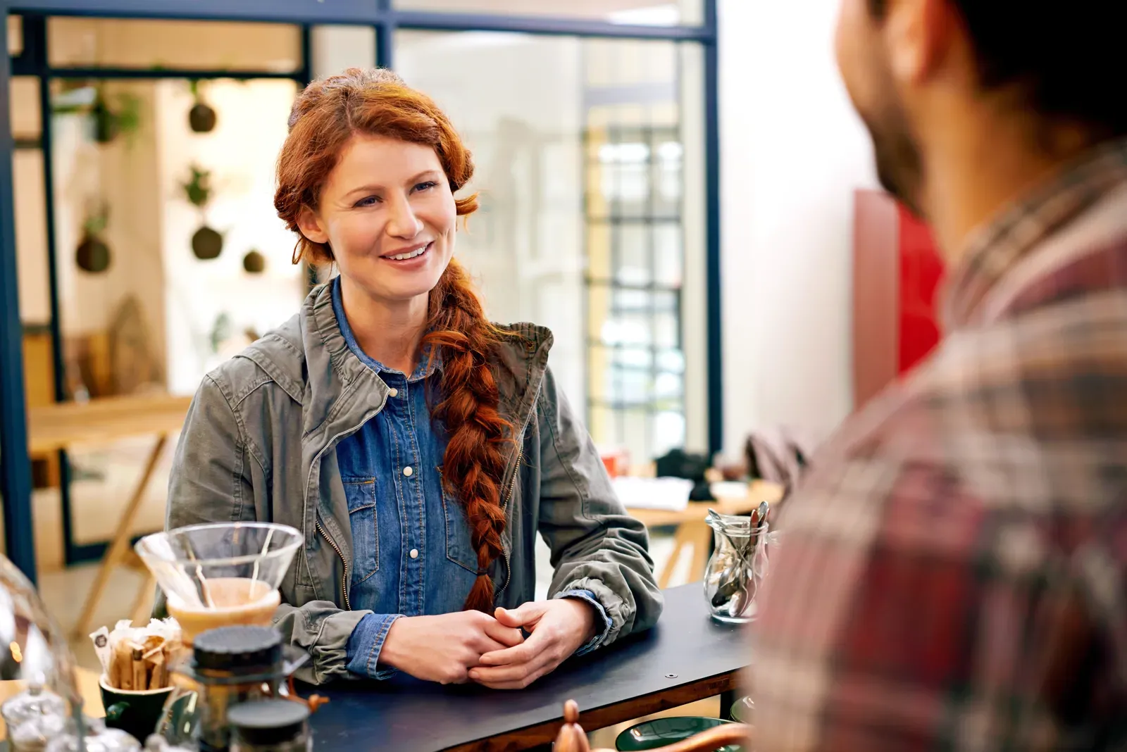 Smiling redhead woman with braided hair leaning on a café counter, conversing with a person across from her.